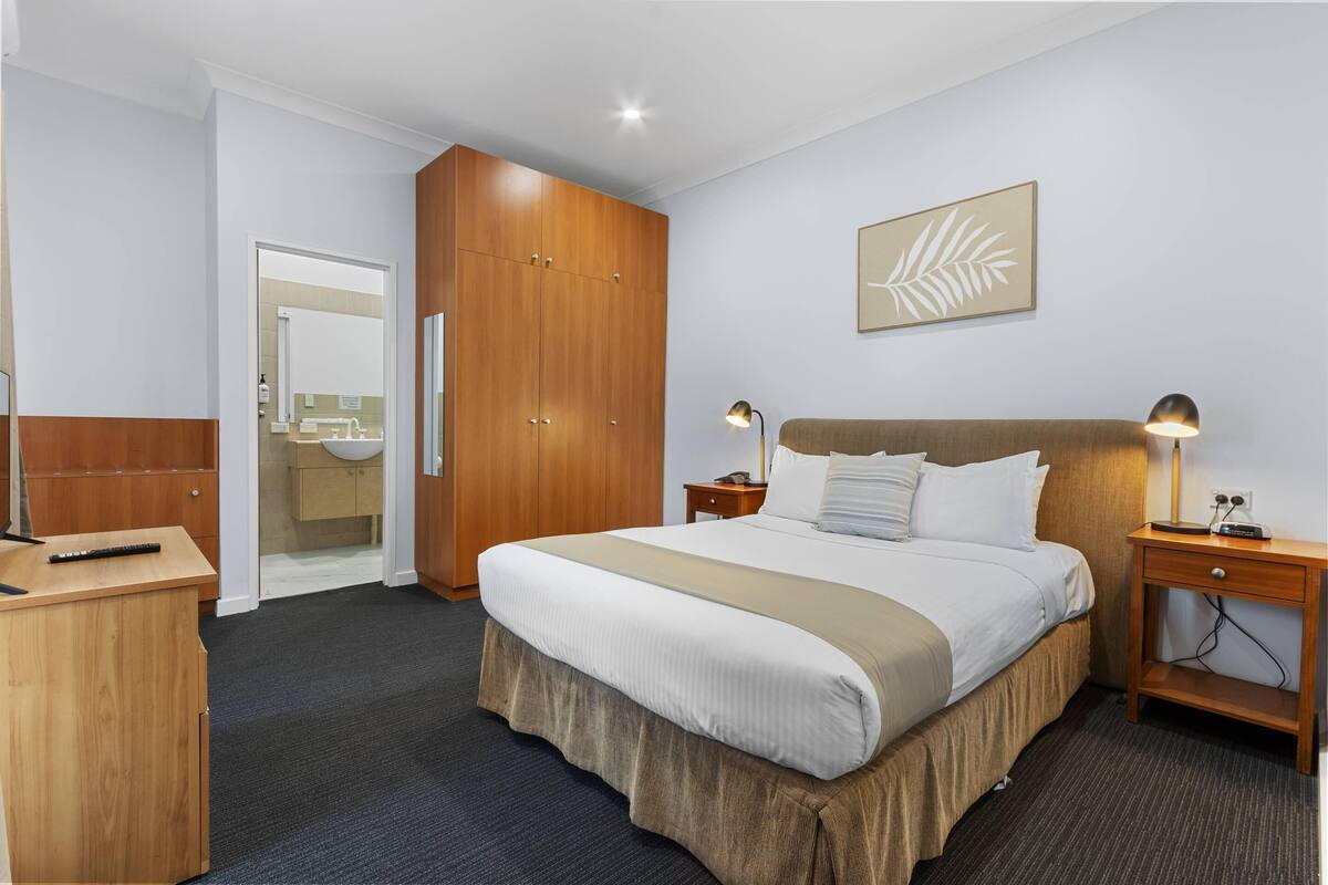 Quarto de hotel com cama, guarda-roupa de madeira, mesas de cabeceira, escrivaninha e banheiro visível por uma porta aberta.
