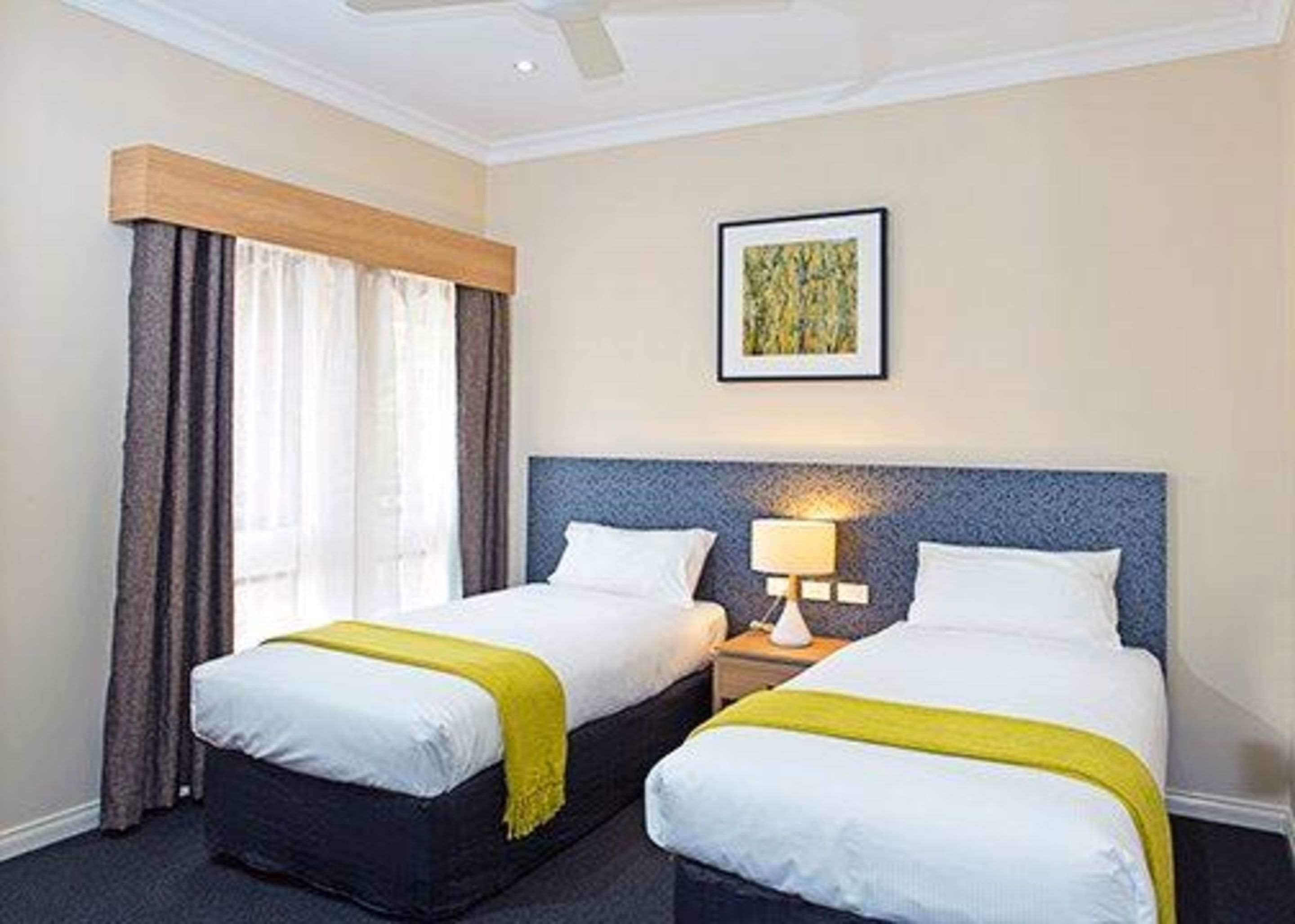 Habitación de hotel con dos camas, ventilador de techo, ventana con cortinas e imagen enmarcada en la pared.