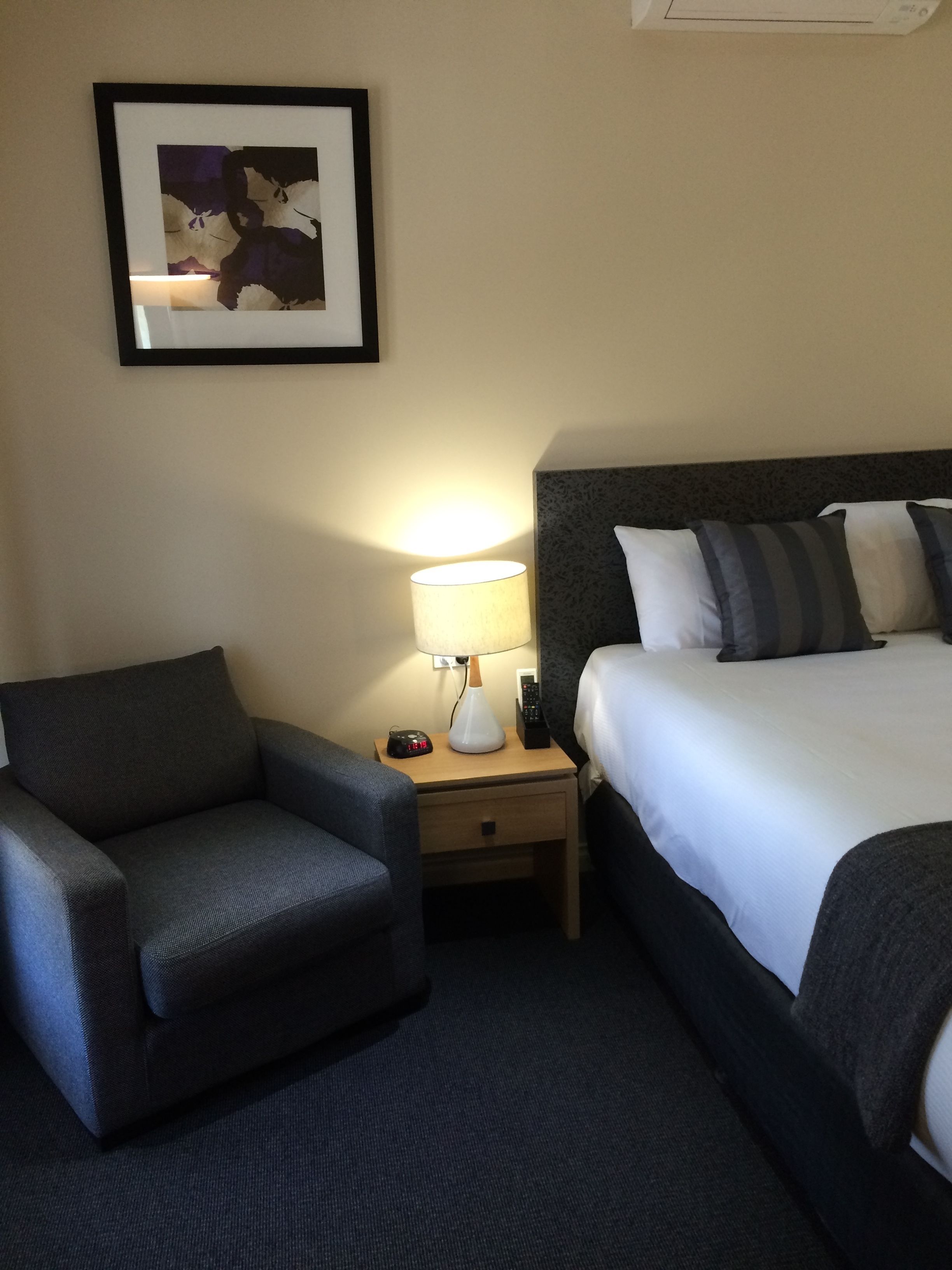 Une chambre d’hôtel avec un lit, une chaise, une table de chevet avec une lampe et un tableau encadré au mur.