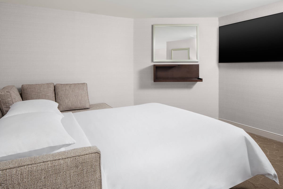 A modern hotel room with a large bed, a flat-screen TV mounted on the wall, a small wooden shelf, and a seating area with a chair and cushions.