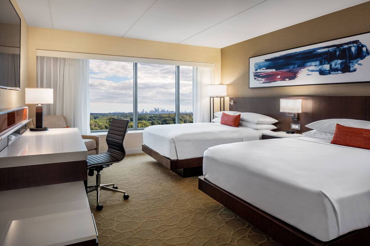 A hotel room with two beds, a desk, a chair, and a large window offering a city view.