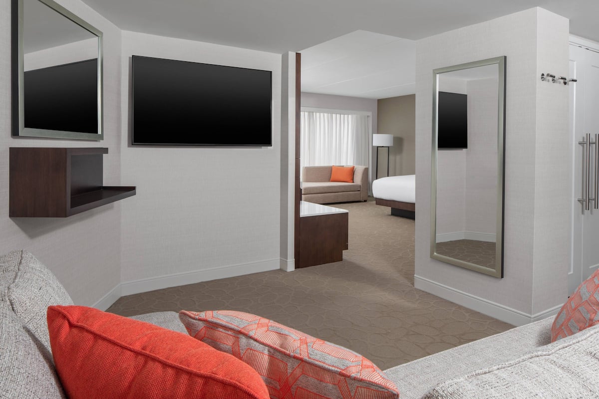 A modern hotel room with a large bed, a seating area with a sofa and armchairs, a TV mounted on the wall, and a large mirror.