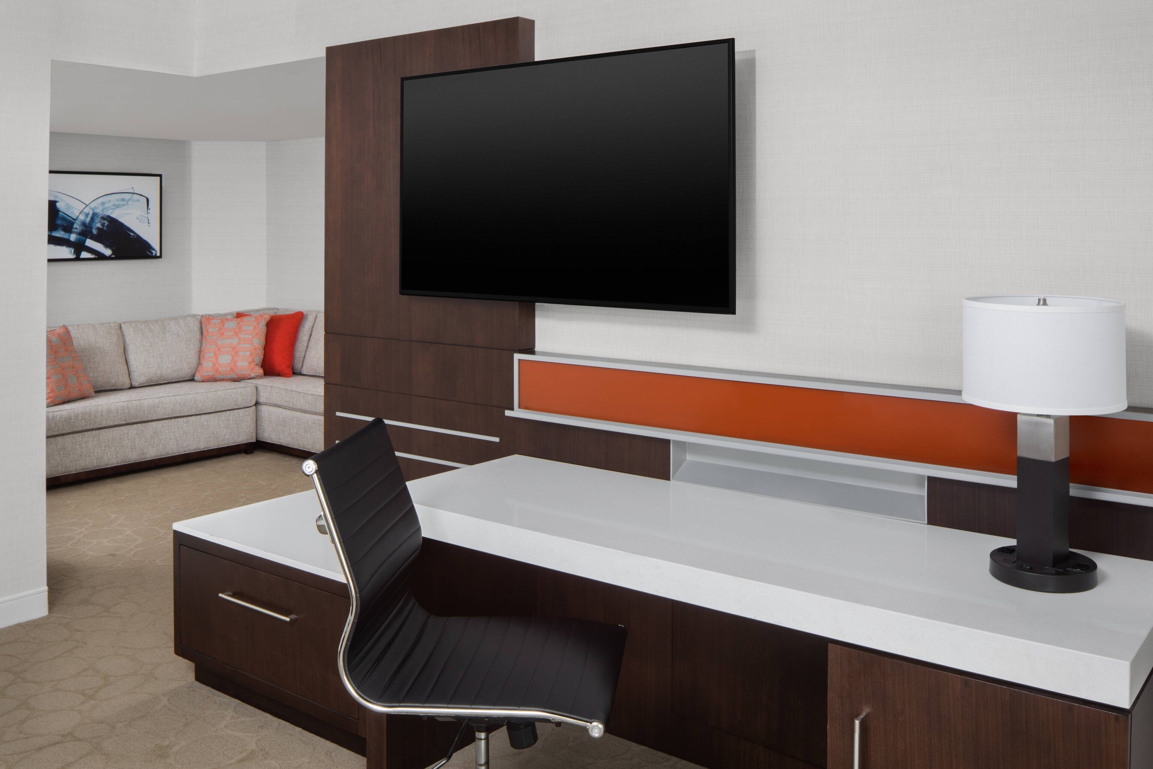 Una habitación de hotel moderna con un televisor de pantalla plana montado en la pared, un escritorio con una silla, un sofá con cojines naranjas y una lámpara.