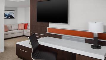 Una habitación de hotel moderna con un televisor de pantalla plana montado en la pared, un escritorio con una silla, un sofá con cojines naranjas y una lámpara.