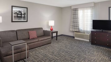 Una habitación de hotel con un sofá gris, una mesita auxiliar con una lámpara, un televisor de pantalla plana, un aparador y un cuadro en la pared.