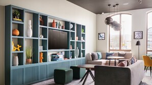 A modern living room with a large TV, teal shelving, green ottomans, and a sofa with multiple cushions. - Holiday Inn Lincoln by IHG (Lincoln)