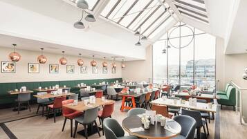 A modern dining area with multiple tables and chairs, large windows, and artwork on the walls.