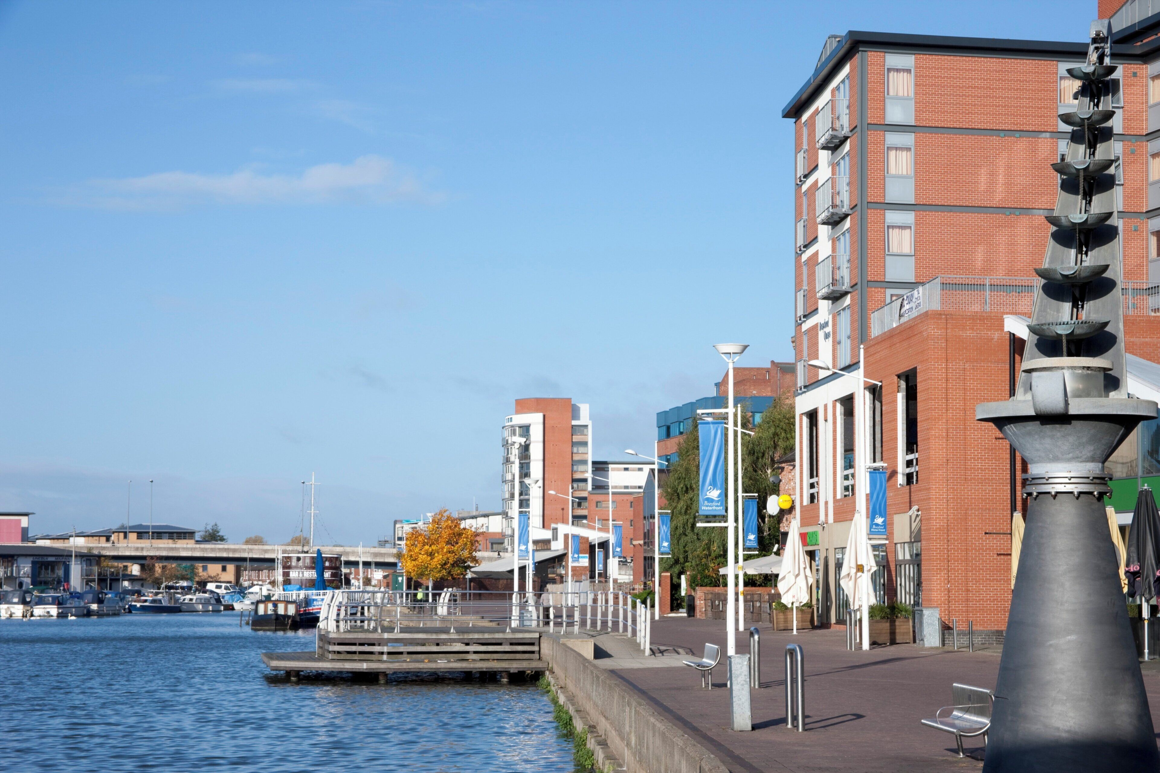A waterfront promenade with a canal, brick buildings, and a modern sculpture.