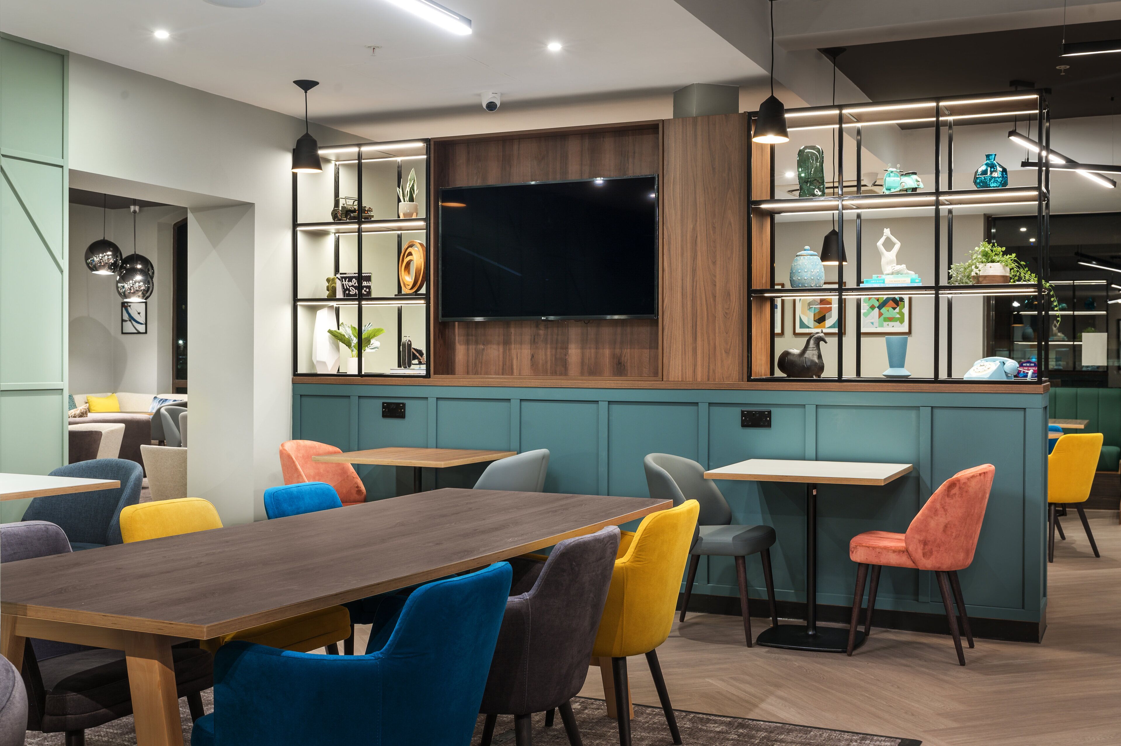 A modern interior with a wooden dining table, colorful chairs, a flat-screen TV, and shelves with decorative items.