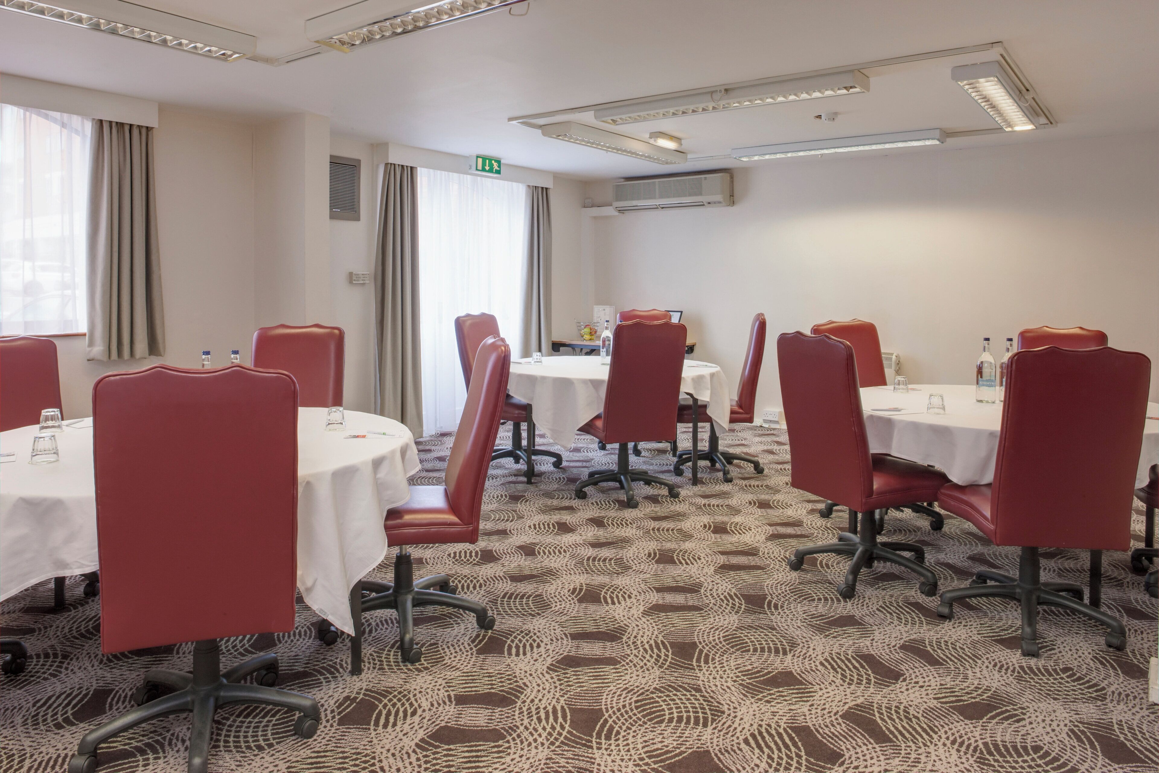 A conference room with round tables and red chairs, air conditioning units on the ceiling, and a patterned carpet.