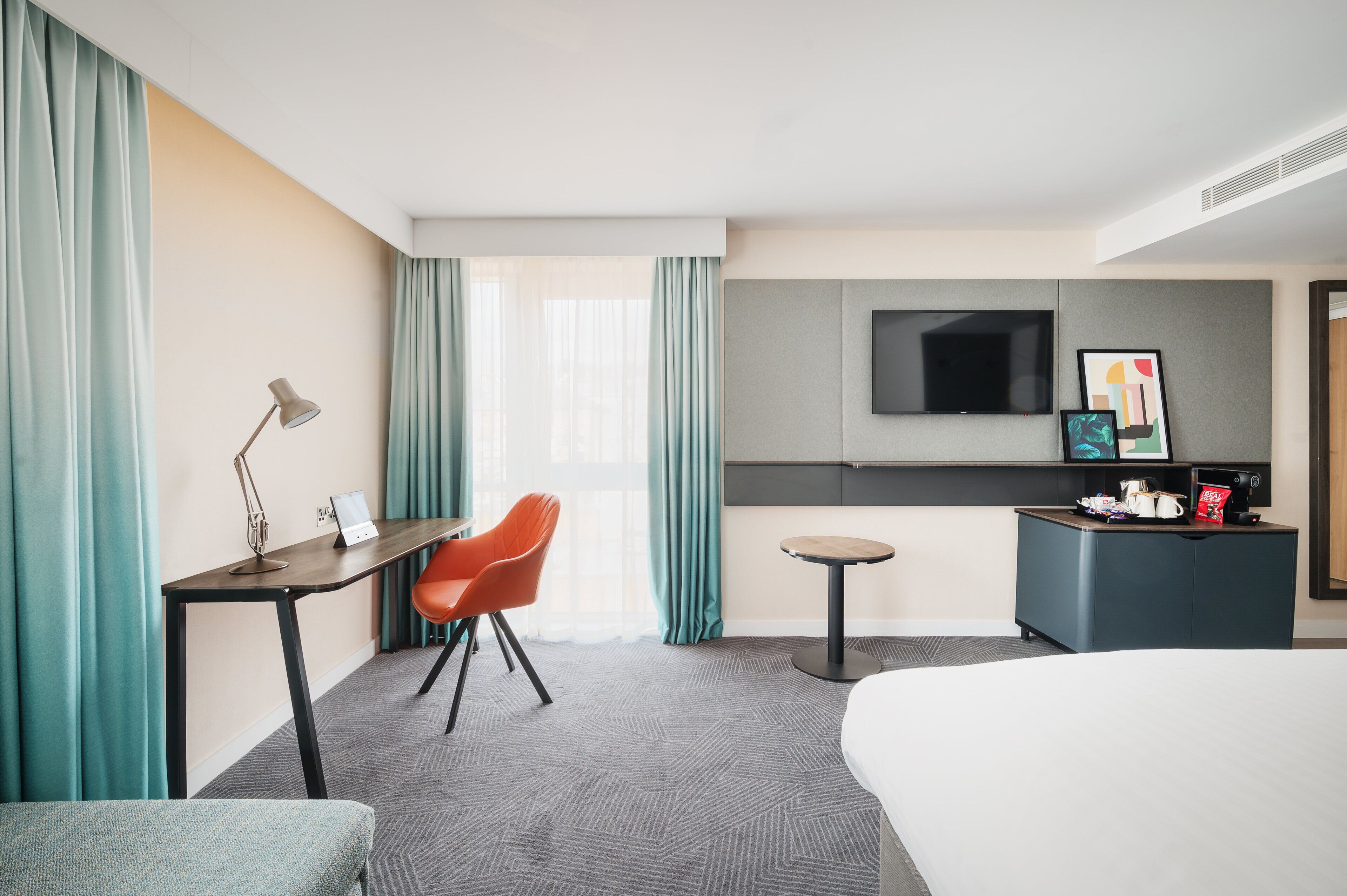 A hotel room with a bed, a desk with a lamp, a chair, a small table, a TV, and a minibar.
