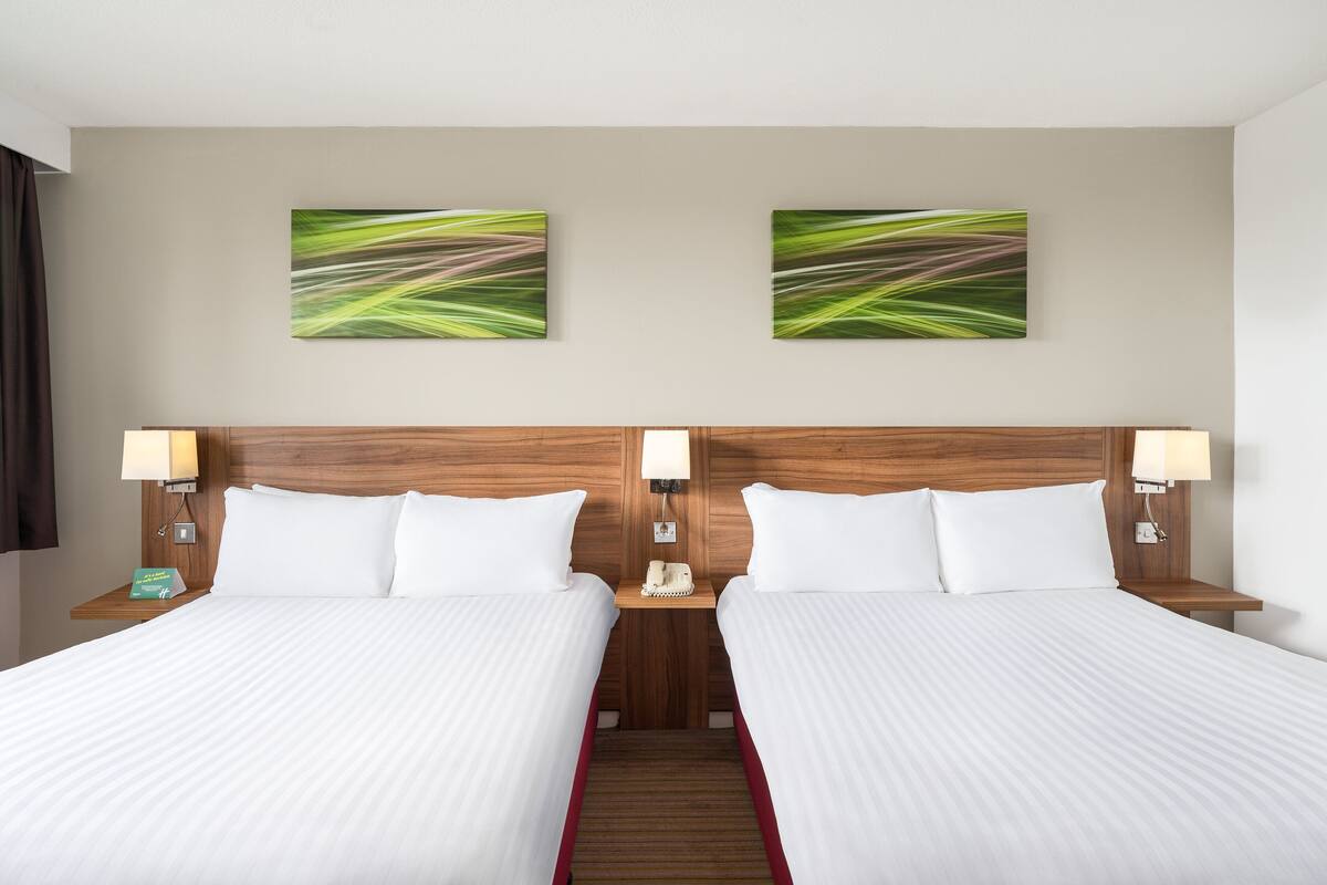 A hotel room with two beds, wooden headboards, white bedding, and two wall-mounted lamps.