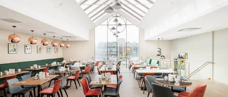 A spacious dining area with multiple tables and chairs, a high ceiling with skylights, and artwork on the walls.
