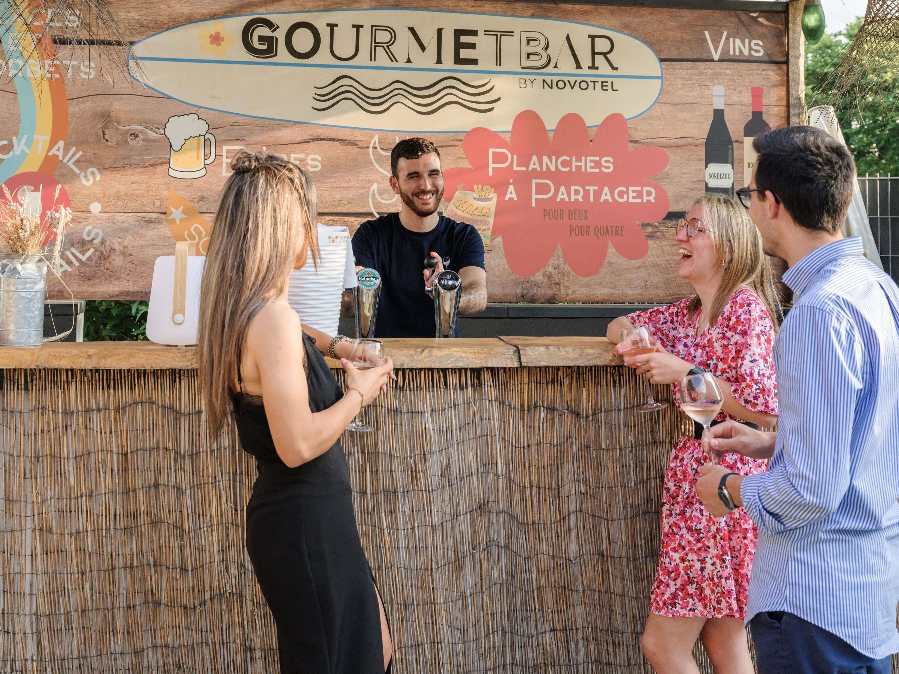 A bar area with a wooden backdrop, a sign reading 'GOURMETBAR BY NOVOTEL,' and a menu offering 'Planches à Partager pour deux' and 'pour quatre.'