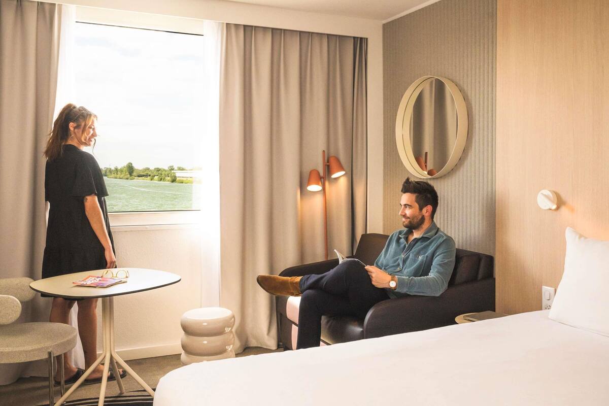 A hotel room with a bed, a round mirror, a desk with a chair, and a view of a body of water through the window.