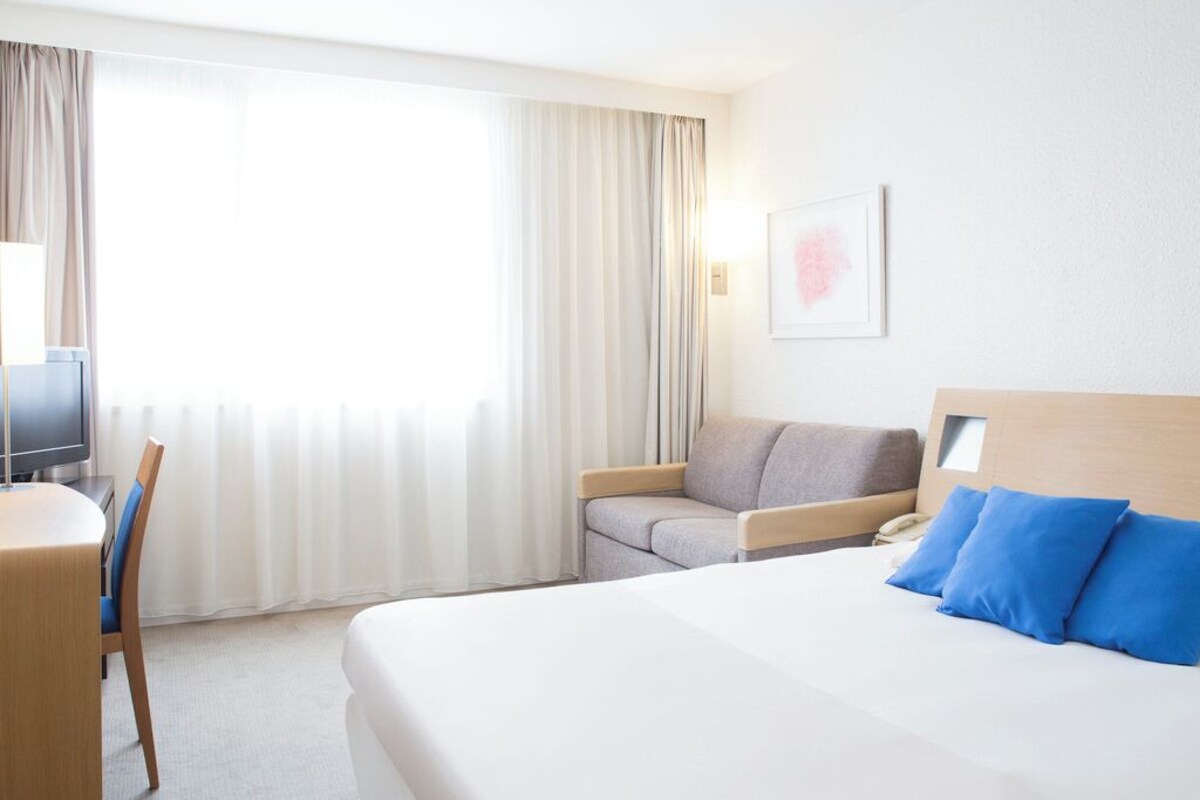 A hotel room with a bed, a desk, a chair, a sofa, and a large window with curtains.