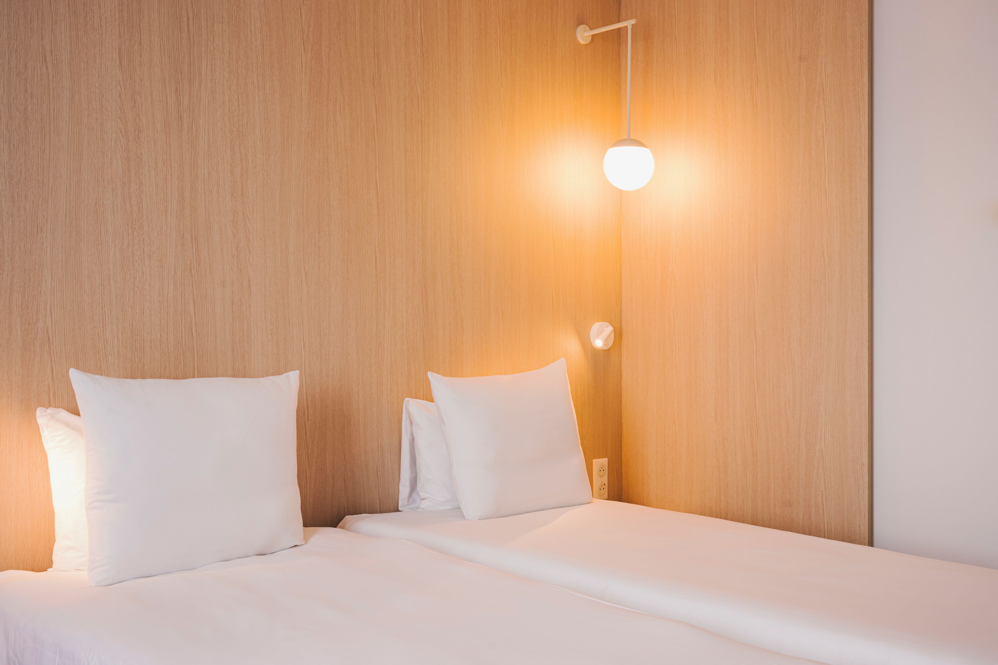 Una cama bien tendida con sábanas y almohadas blancas, cabecero de madera y una lámpara fijada a la pared.