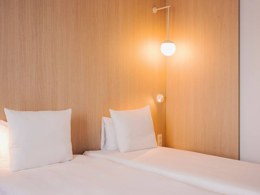 Una cama bien tendida con sábanas y almohadas blancas, cabecero de madera y una lámpara fijada a la pared.
