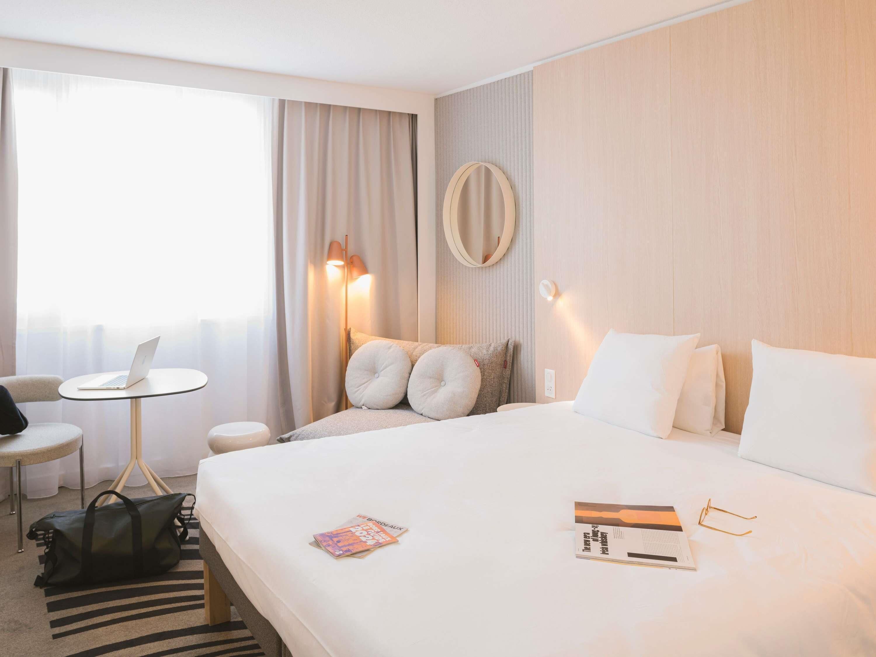 A hotel room with a bed, a desk with a laptop, a chair, a lamp, and a window with curtains.