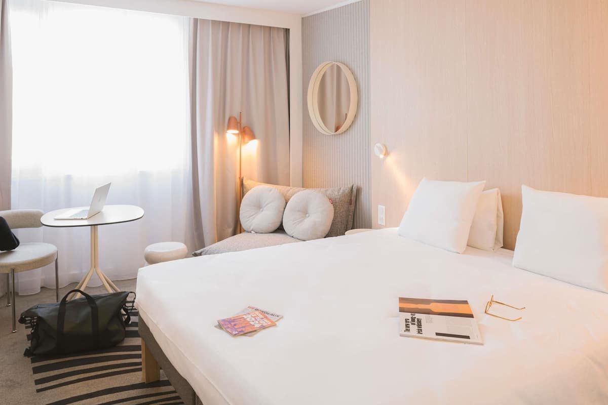 A hotel room with a bed, a desk with a laptop, a chair, a lamp, and a window with curtains.