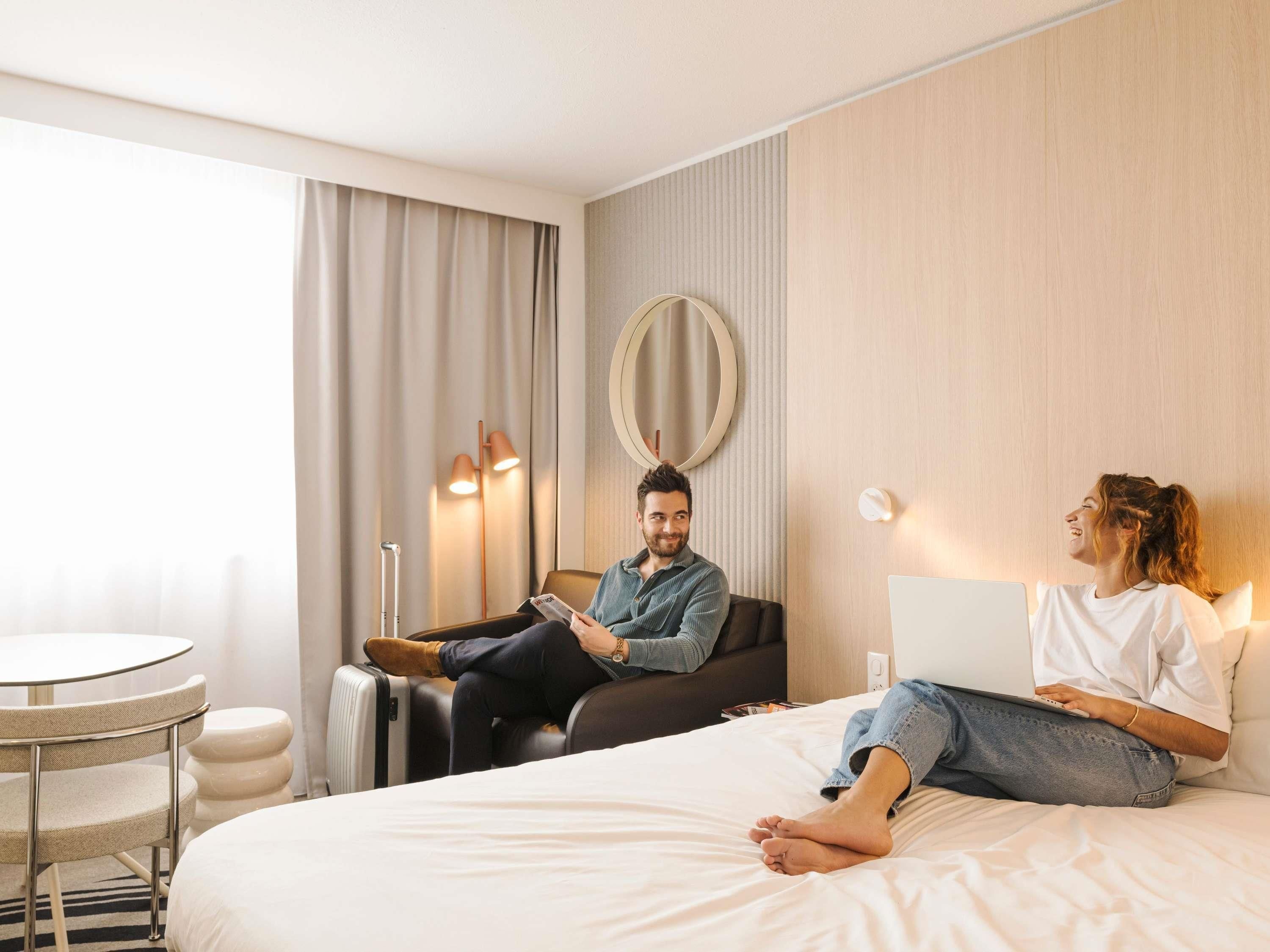 A hotel room with a bed, a round mirror, a standing lamp, and a laptop on the bed.