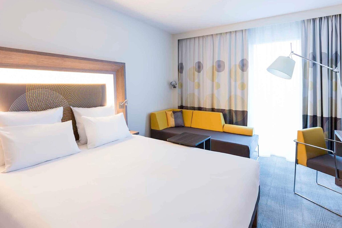 A hotel room with a large bed, a sofa, a small table, and a chair.