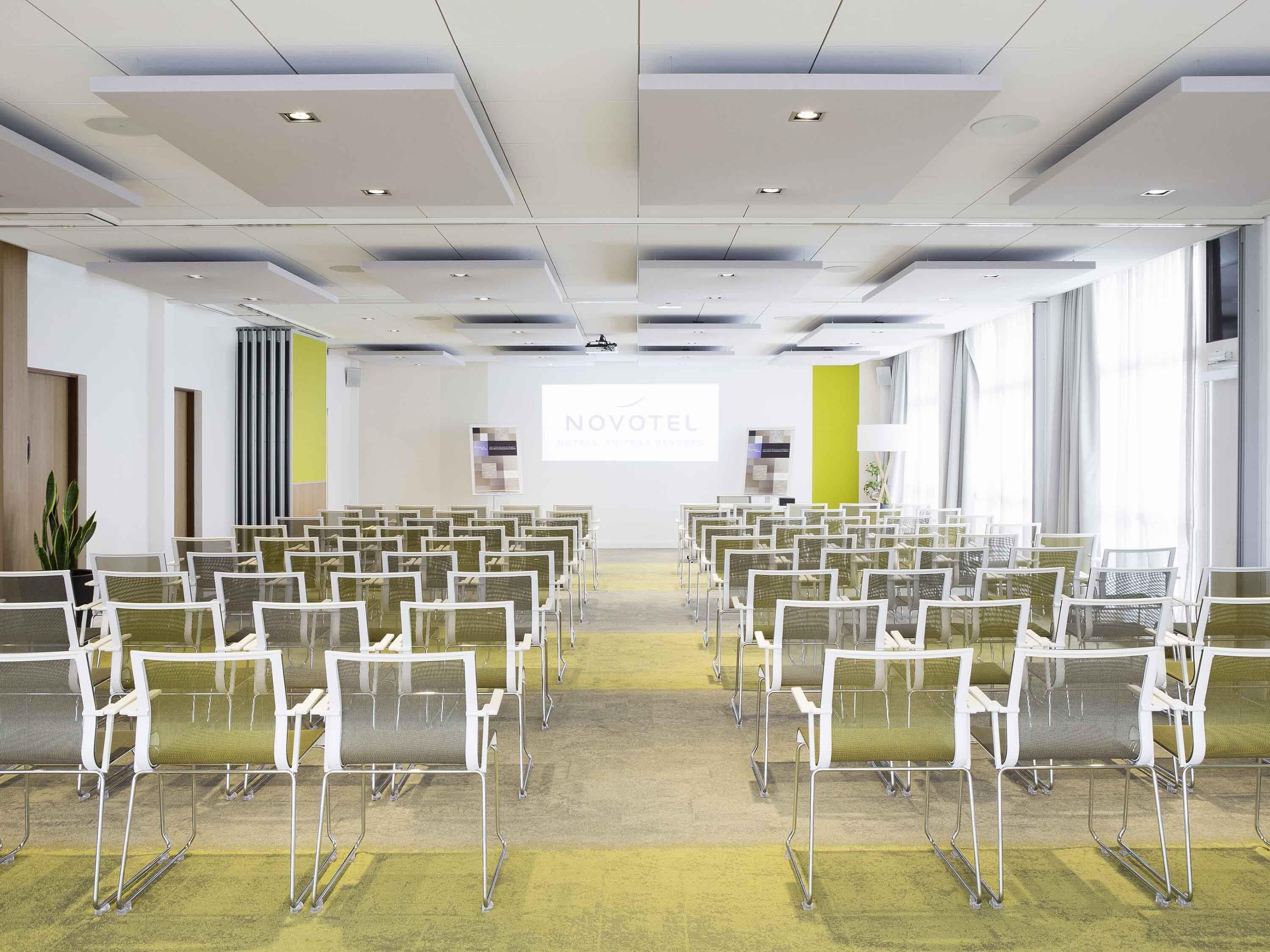 Un salón de conferencias con filas de sillas, una pantalla de proyector y un letrero con la marca NOVOTEL.
