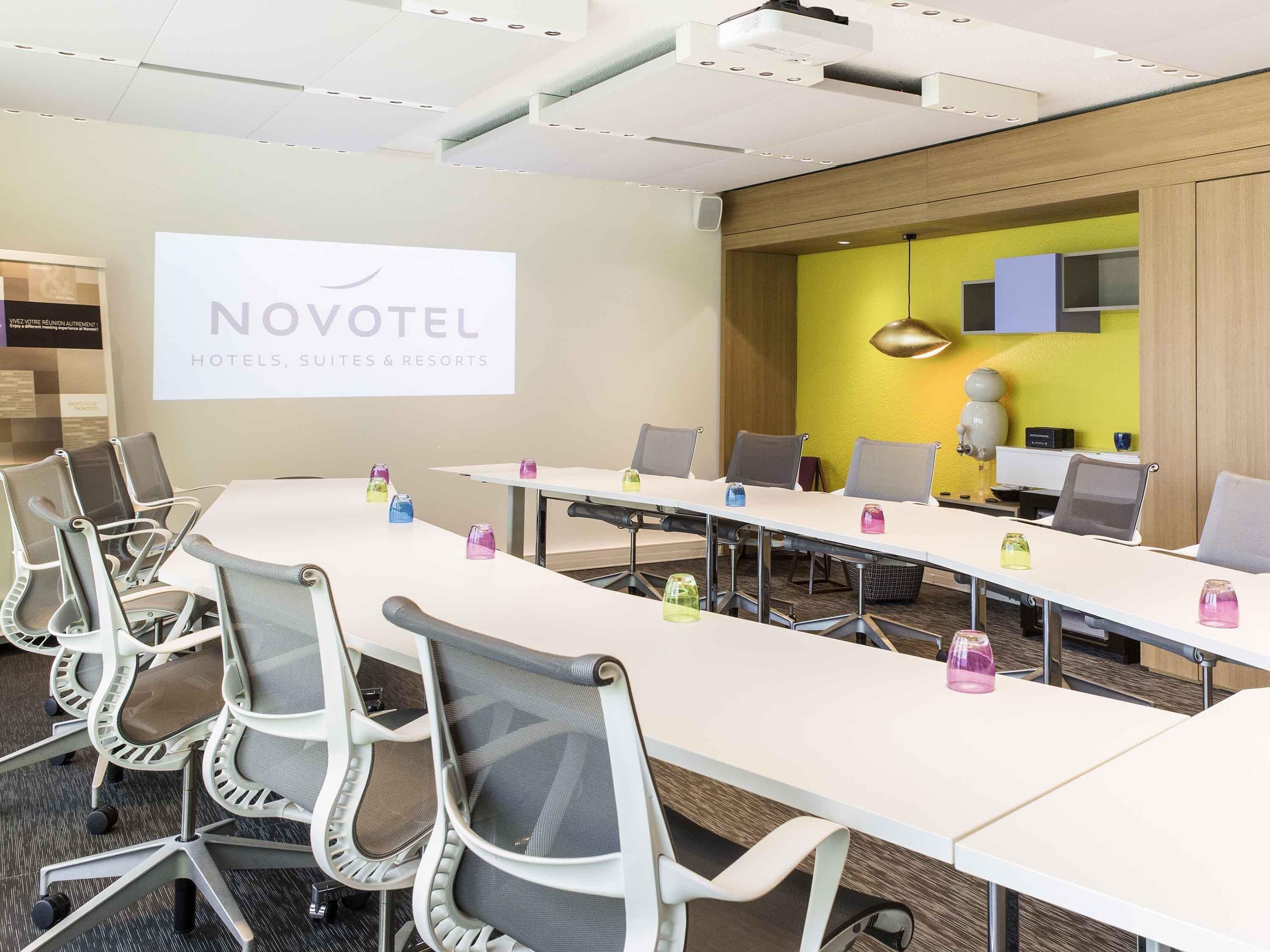 Une salle de conférence équipée d’une longue table, de chaises et d’un écran de projection sur lequel est affiché le texte « NOVOTEL HOTELS, SUITES & RESORTS ».