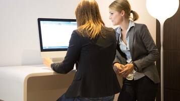 Dos personas interactúan con un monitor de computadora en una habitación con un escritorio y un taburete blancos.