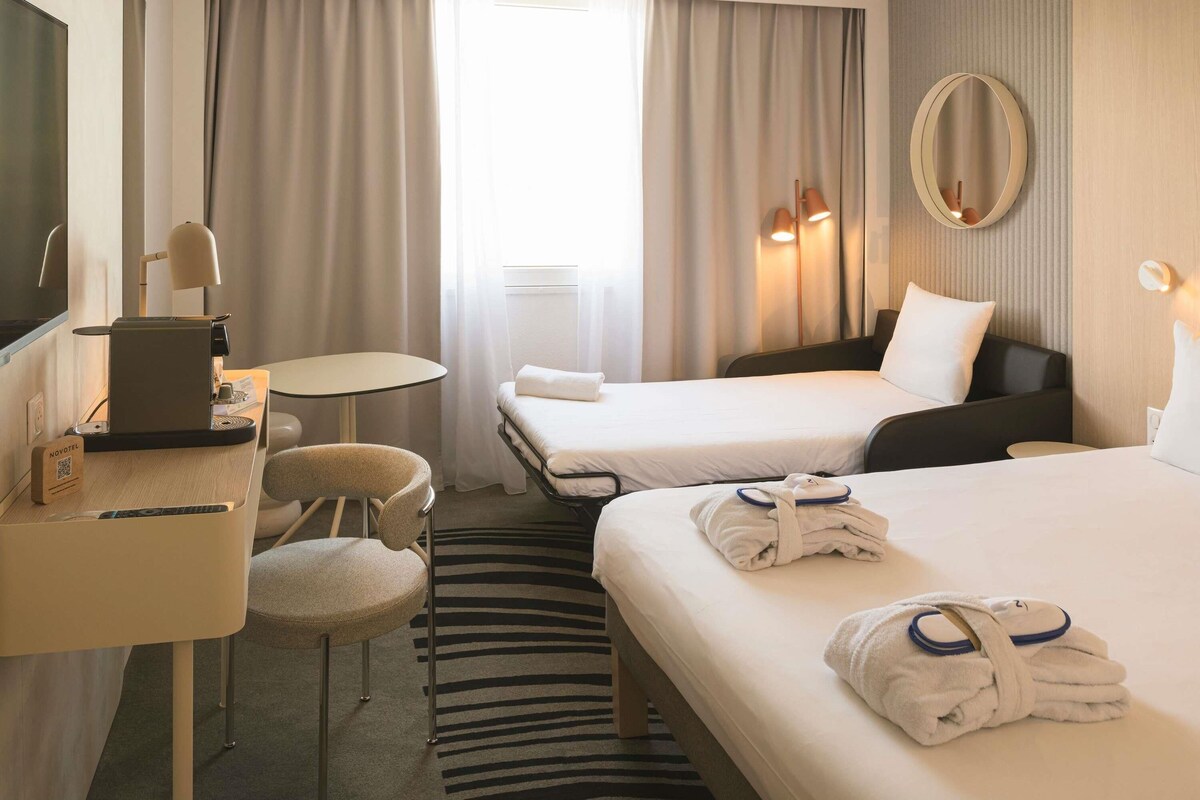 A hotel room with two beds, a desk with a laptop, a chair, a small table, a mirror, and a window with curtains.