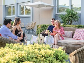 A group of people seated on a wicker sofa outdoors with a table between them, surrounded by greenery and a building in the background. - Novotel Bordeaux Lac (Bordeaux)