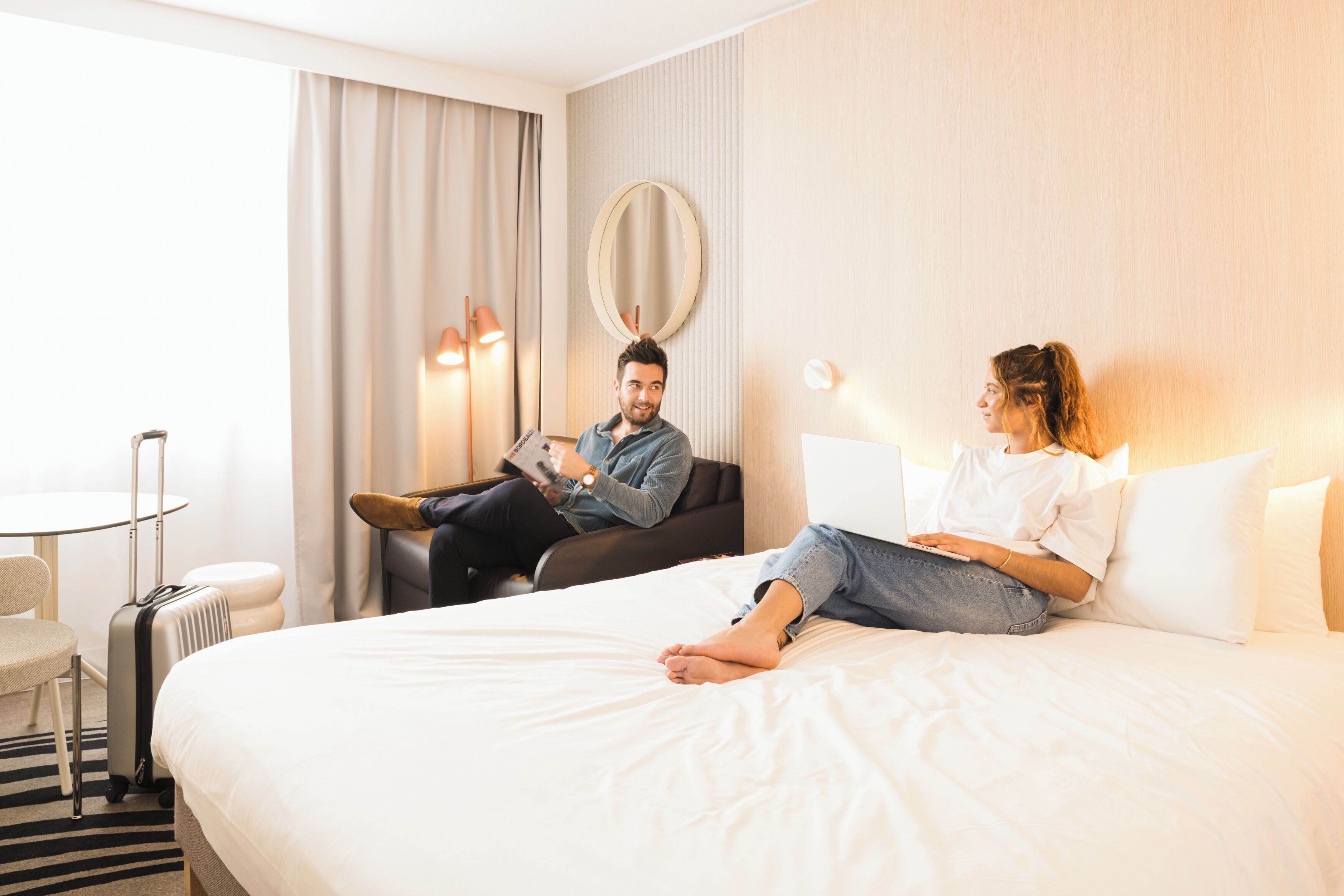 A hotel room with a bed, a round mirror, a table, and a chair.
