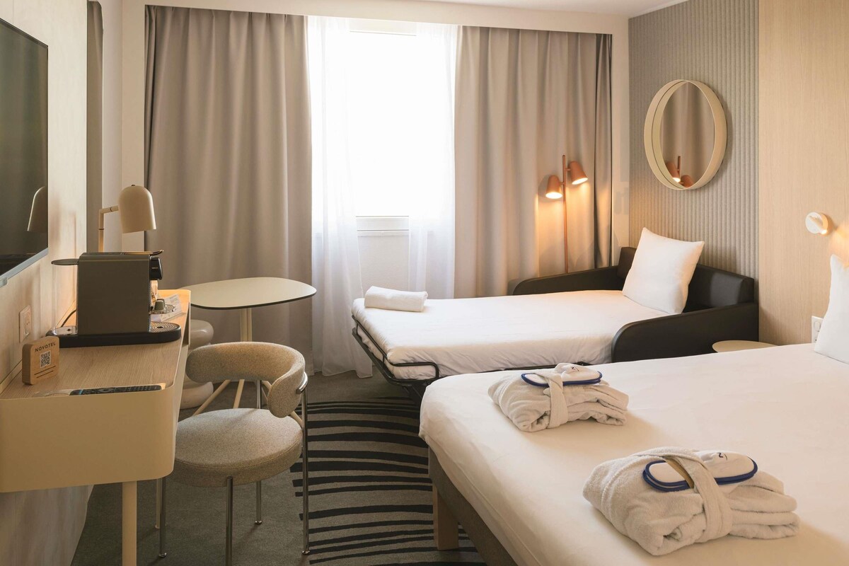 A hotel room with two beds, a desk with a laptop, a chair, a small table, a mirror, and a window with curtains.