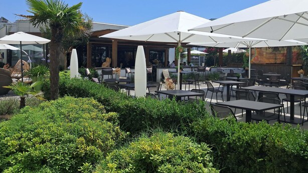 An outdoor cafe with tables and umbrellas, surrounded by greenery and a building in the background. - Novotel Bordeaux Lac (Bordeaux)