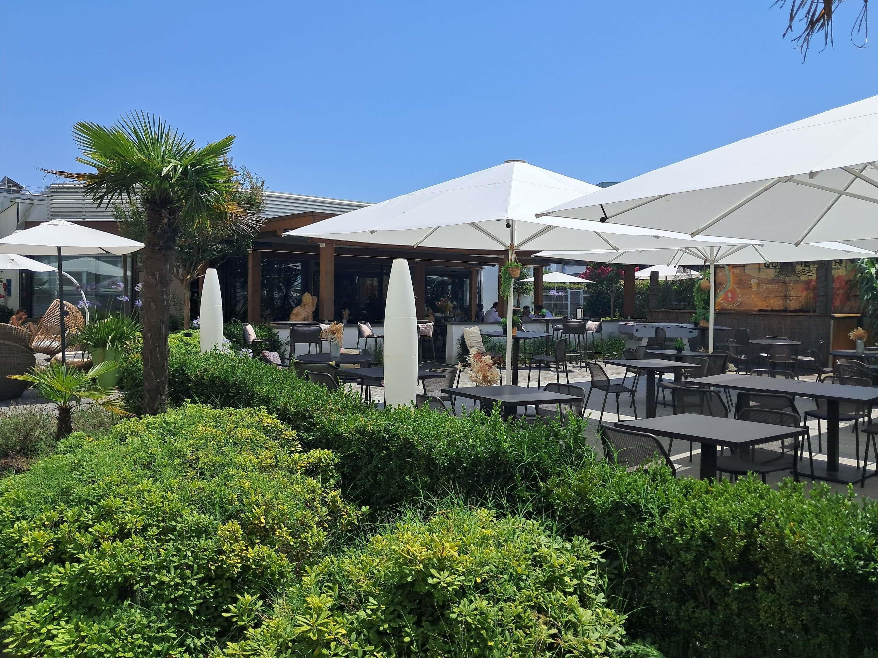 An outdoor seating area with tables and umbrellas, surrounded by greenery and a building in the background.