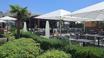An outdoor seating area with tables and umbrellas, surrounded by greenery and a building in the background.