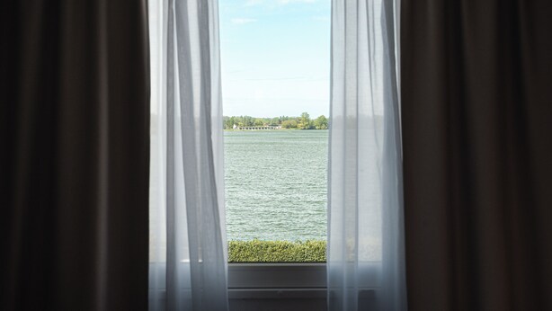 Vista a través de una ventana con cortinas translúcidas, que muestra un cuerpo de agua tranquilo y una costa distante.