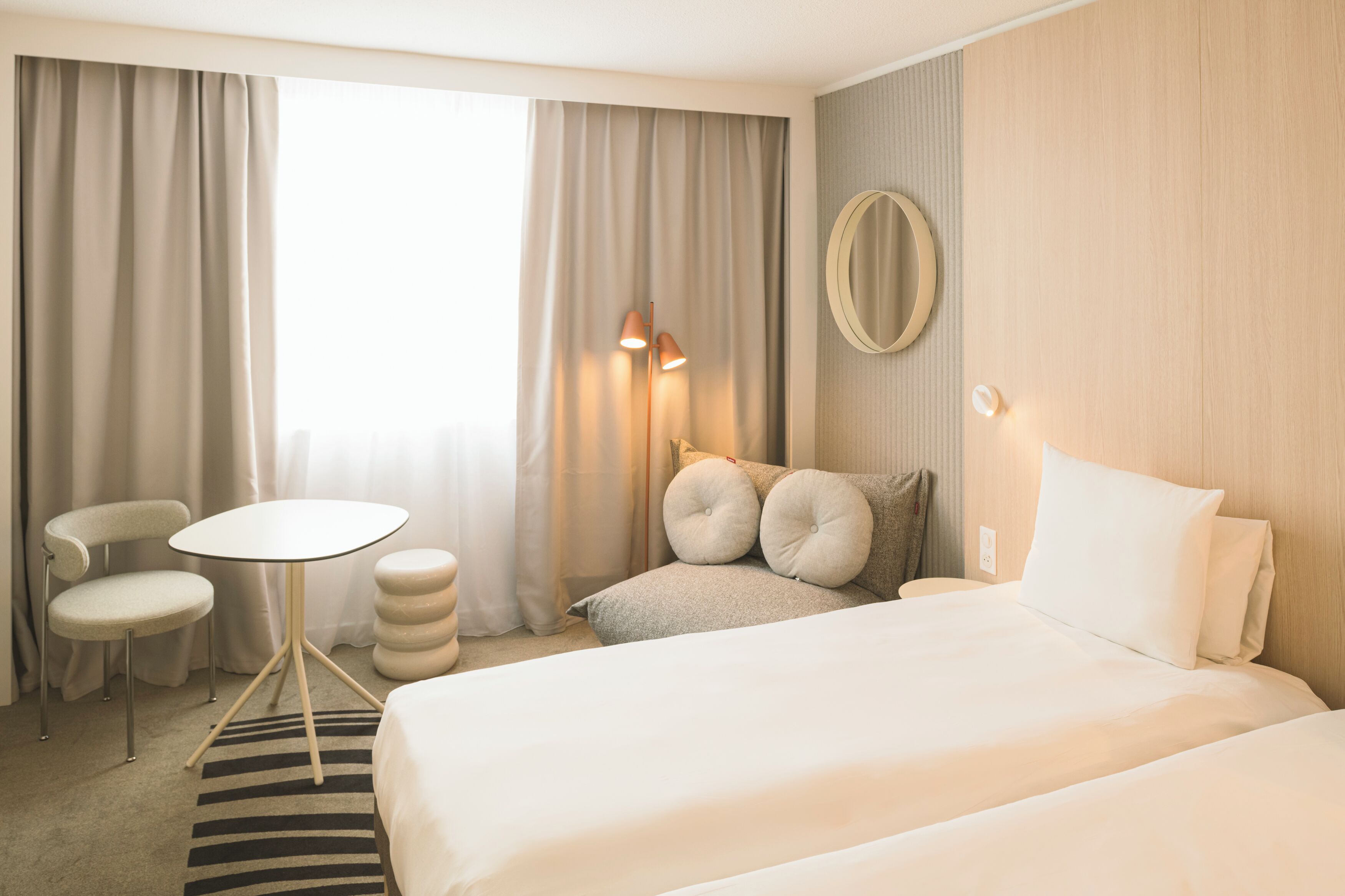 Habitación de hotel con cama, un espejo redondo, una mesita pequeña, dos sillas y una alfombra a rayas.
