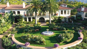 Un edificio grande y blanco con un patio central que cuenta con una fuente, rodeado de exuberantes jardines y palmeras.