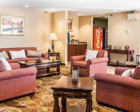 A hotel room with a patterned red sofa set, a wooden coffee table, a lamp, a Coca-Cola refrigerator, and a framed picture on the wall.
