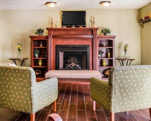 A living room with a fireplace, two armchairs, a television above the fireplace, and shelves with various items.