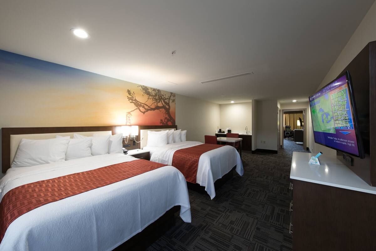 A hotel room with two beds, a desk, a TV, and a wall with a tree painting.
