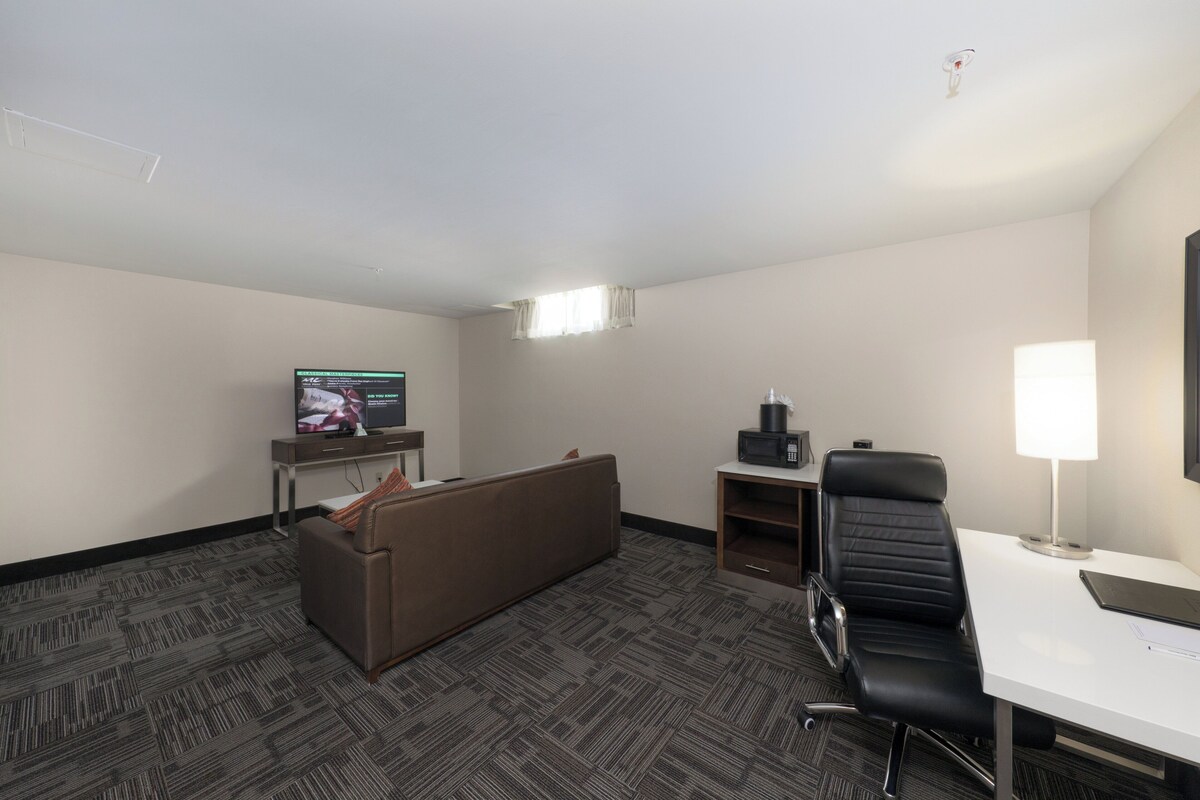A hotel room with a sofa, a desk, a chair, a television, and a lamp.