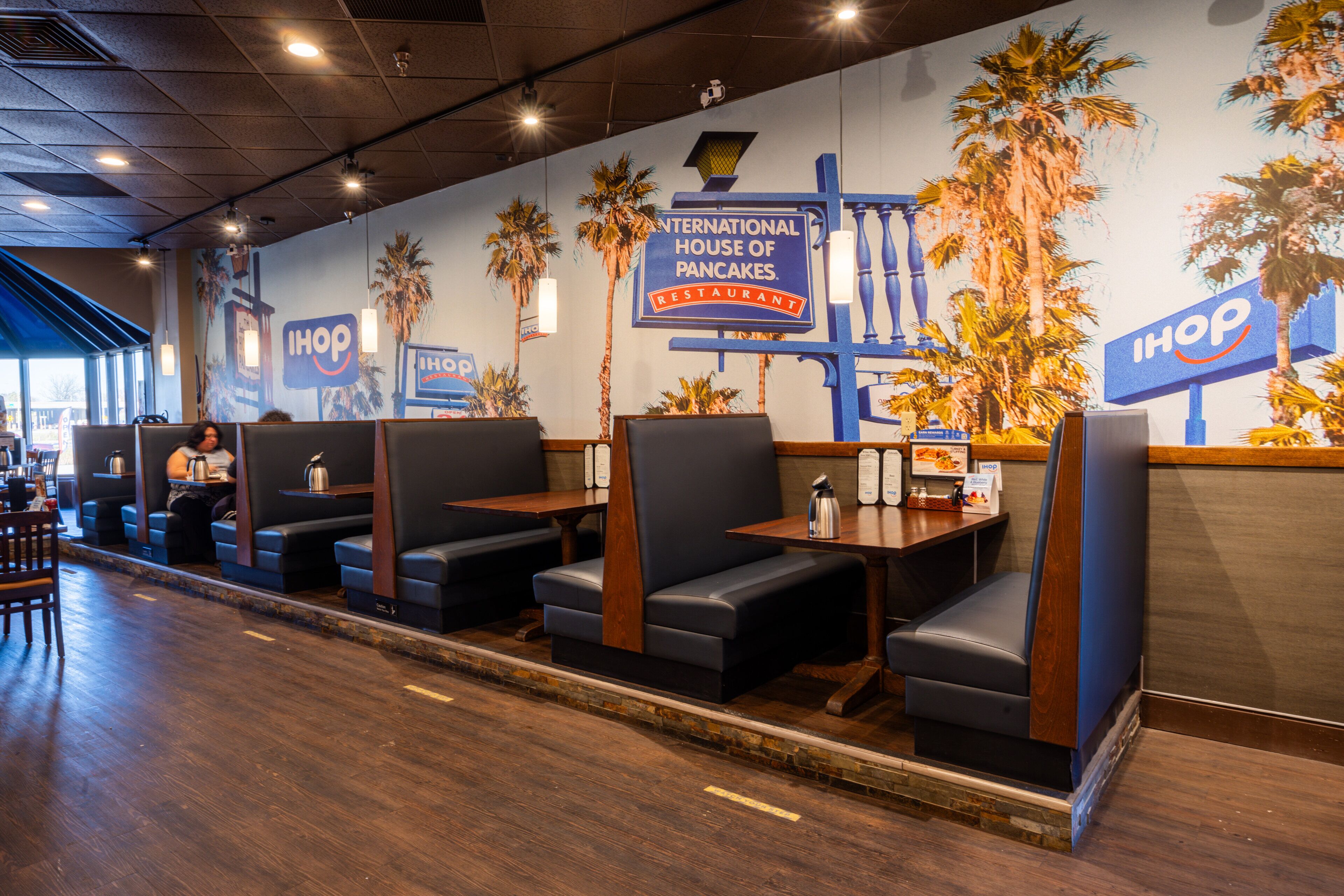L’intérieur d’un restaurant avec des banquettes, des tables en bois et une fresque représentant des palmiers, ainsi qu’une enseigne de la chaîne International House of Pancakes.