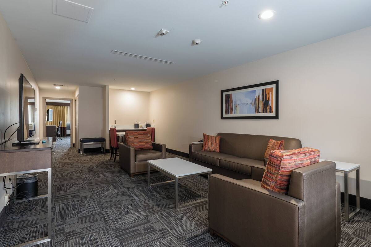 A hotel room with a sofa, armchair, and a small table. There is a TV on a stand and a window with curtains.