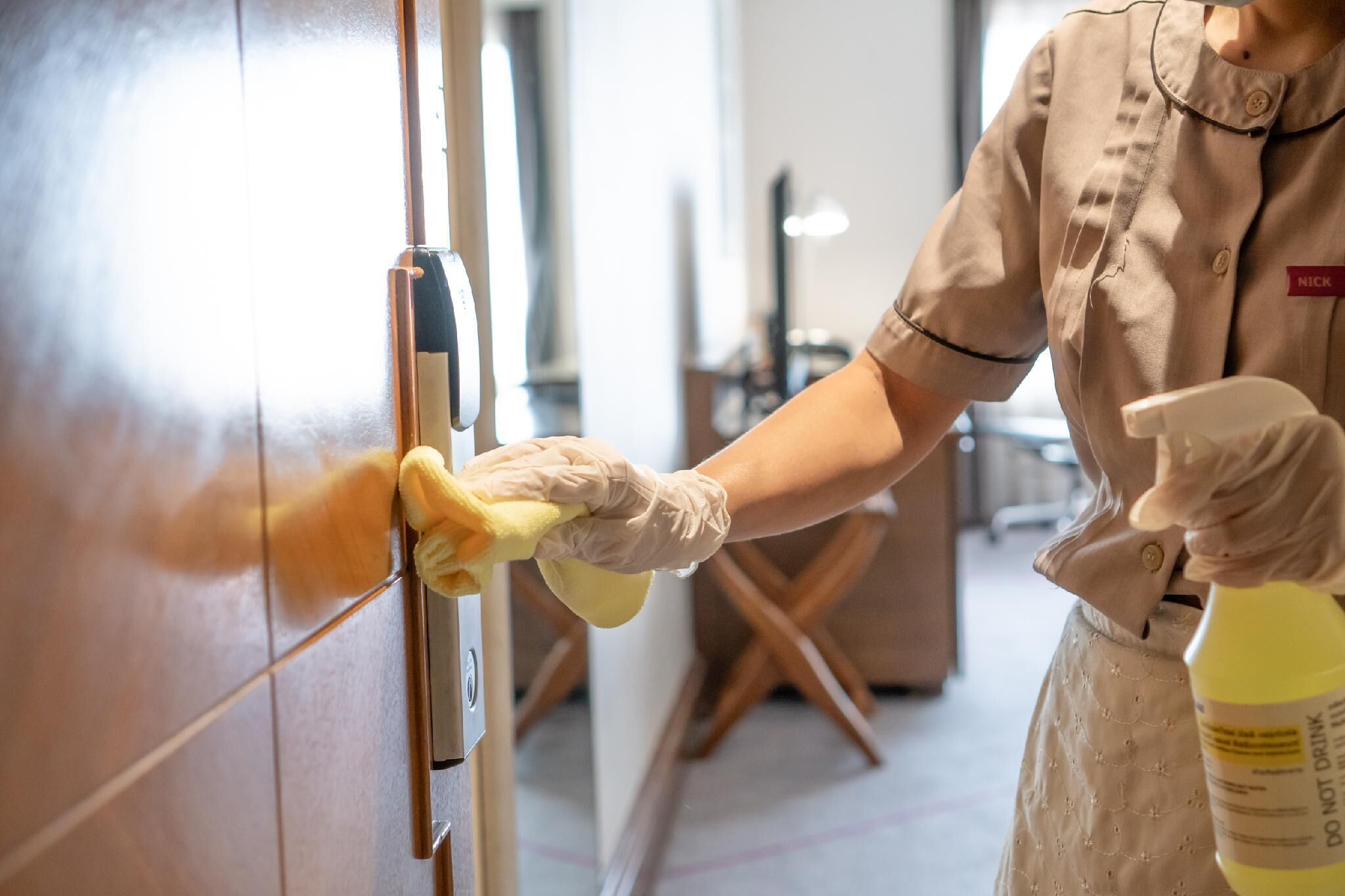 A person cleaning a door handle with a yellow cloth and a spray bottle labeled "DO NOT DRINK".