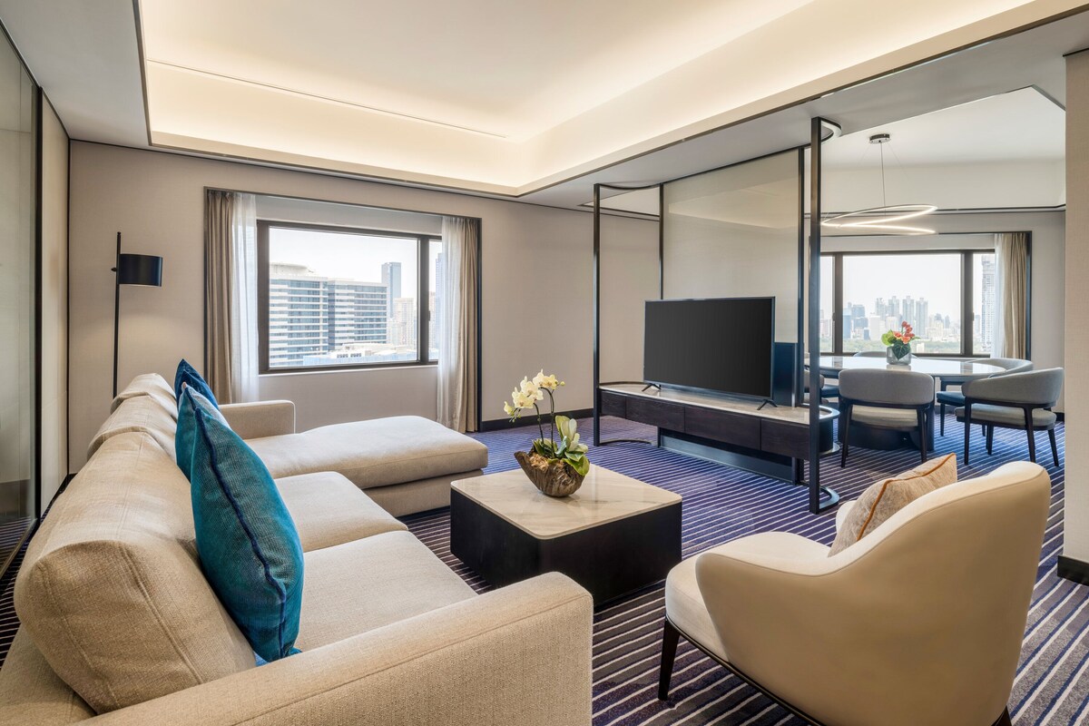 Una habitación de hotel moderna con sofá, sillón, mesa de centro y televisión. Cuenta con un área de comedor con mesa y sillas, y un amplio ventanal con vistas a la ciudad.