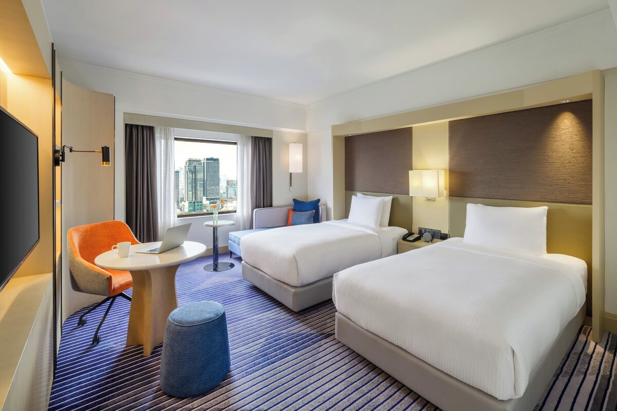 Habitación de hotel con dos camas, un escritorio, una silla, una mesita y vistas a la ciudad.