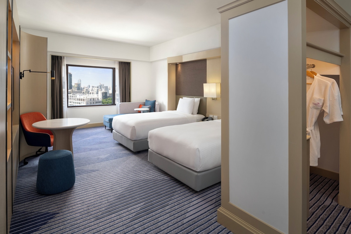 Habitación de hotel con dos camas, una mesita, una silla y vistas a la ciudad.