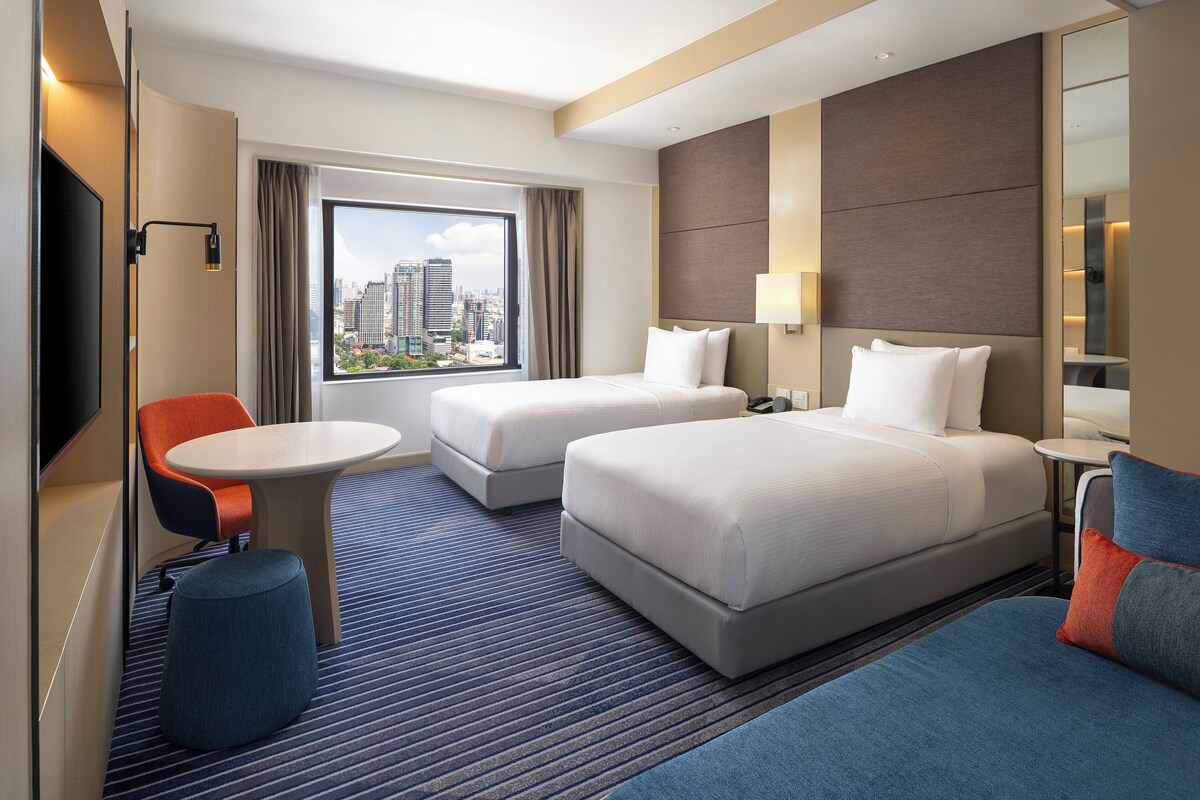Habitación de hotel con dos camas, un escritorio, una silla y un ventanal con vistas a la ciudad.